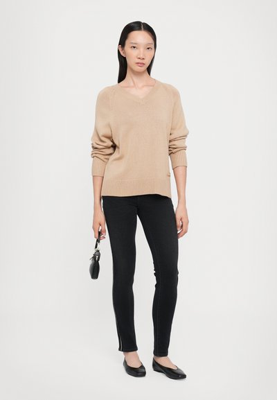 Jersey beige de punto con escote en V, puños acanalados y dobladillo. Combinado con unos jeans negros ajustados que presentan un detalle en la costura lateral. Zapatos planos negros.