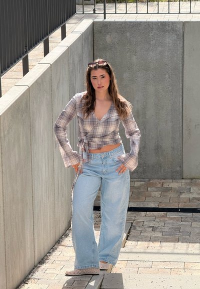 Joven mujer de pie al aire libre frente a paredes de hormigón, vistiendo una camisa cruzada de cuadros, vaqueros de pierna ancha en azul claro y gafas de sol en la cabeza.