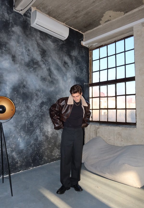 Uomo con giacca in pelle marrone con montone e pantaloni neri si trova in una stanza industriale con una grande finestra, una lampada su treppiede e una poltrona a sacco.