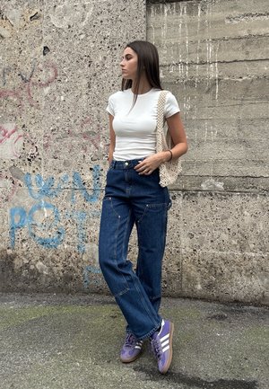 Weißes, eng anliegendes T-Shirt, hoch taillierte blaue Jeans und lila Sneaker mit weißen Streifen; trägt eine strukturierte, gewebte Tasche.