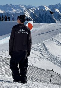 Černá dlouhá rukávová košile s bílým logem, černé kalhoty a černá čepice. Sníh pokrytá horská scenérie s lyžaři a sněhovým skokem.