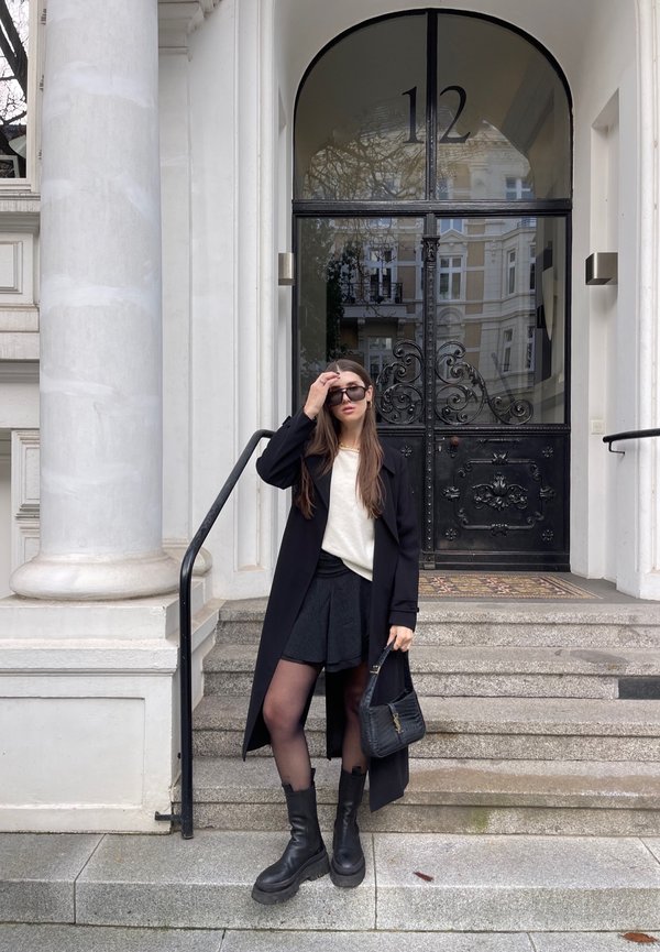 Black long coat, white sweater, and black skirt with tights. Black combat boots and handbag. Minimalist black accessories. A column and door in the background.