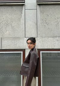 Braune, kurze Lederjacke mit strukturiertem Design, ausgestattet mit einer Klappentasche und Knopfverschluss, vor einer strukturierten grauen Wand.