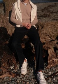 Άτομο κάθεται σε μεγάλο ξεβρασμένο ξύλο κοντά στο νερό, φοράει μπεζ ζιπ-σουίτερ, λευκά παπούτσια, μαύρο παντελόνι και λευκό μπουφάν draped στους ώμους.