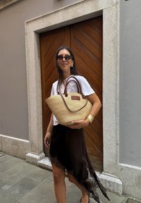 Beige szőtt szalma táska barna műbőr füllel és egy kis címkével; fehér felsővel és sötétbarna aszimmetrikus szoknya viseli.
