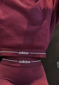 Ensemble de sport bordeaux comprenant un haut ajusté et une ceinture assortie. Logo "adidas" blanc et bandes d'accent visibles. Texture lisse.