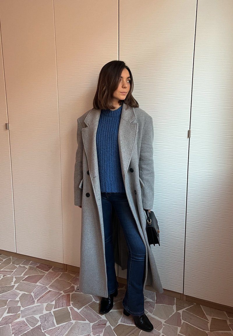 Grey wool coat with black buttons, layered over a blue knitted jumper, paired with dark flared jeans and black ankle boots. Small black handbag.
