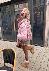 Lyserød puffejakke, ternet mininederdel, brune ankelstøvler, lyserøde ørevarmere med et mønster og en kop i hånden, stående på en belagt fortov.