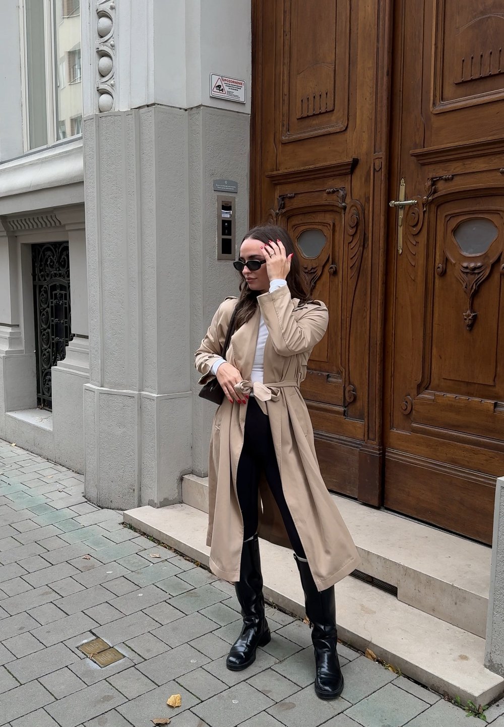 Beige trenchcoat med belte i midjen, over en hvit topp og svarte leggings. Sorte knelange støvler. Står på en steinlagt gate.