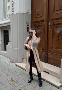 Beige trenchcoat med belte i midjen, over en hvit topp og svarte leggings. Sorte knelange støvler. Står på en steinlagt gate.