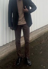Homme debout à l'extérieur portant un blazer noir, un pull rayé marron, un pantalon marron et des chaussures habillées noires près d'un mur blanc ondulé.