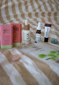Tre blush stick Pixi colorati, un siero trasparente e un tubo di balsamo per labbra su una superficie a righe con accenti decorativi a forma di conchiglia.