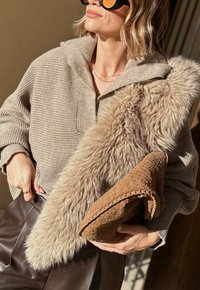 Femme portant un pull surdimensionné en maille beige, un pantalon en cuir marron, tenant une pochette tressée beige et portant un stole en fausse fourrure beige ainsi que des lunettes de soleil.