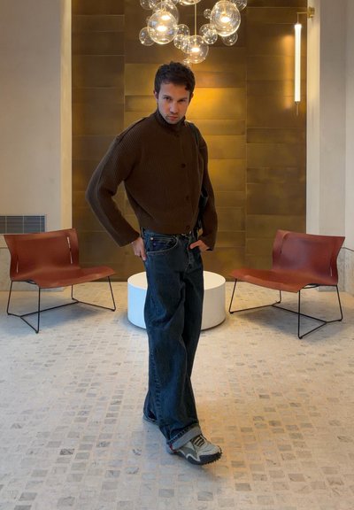 Hombre con suéter marrón y jeans de pie entre dos sillas marrones en una habitación moderna con suelo de baldosas y lámpara de araña de globo de cristal en el techo.