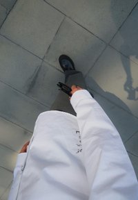Camisa branca de mangas longas com texto preto, calças cinzentas e sapatos pretos, segurando óculos de sol, vista de cima enquanto caminha sobre um pavimento cinzento.