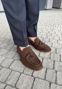 Osoba ubrana w ciemnoniebieskie spodnie i brązowe zamszowe mokasyny z frędzlami stojąca na szarej kostce brukowej.