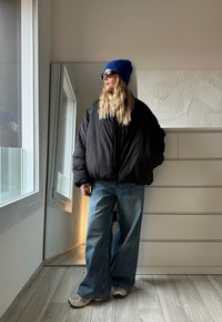 Casaco puffer preto acolchoado, jeans azuis largos, ténis cinzentos e brancos, gorro azul de malha e óculos de sol; em ambiente interno com fundo espelhado.