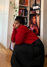 Κόκκινος πλεκτός σφιχτός πουλόβερ με ραβδωτή υφή, συνδυασμένος με μια μαύρη φούστα. Το φόντο περιλαμβάνει ράφια γεμάτα βιβλία και κορνιζαρισμένα έργα τέχνης. Εσωτερικός χώρος με ξύλινο πάτωμα.