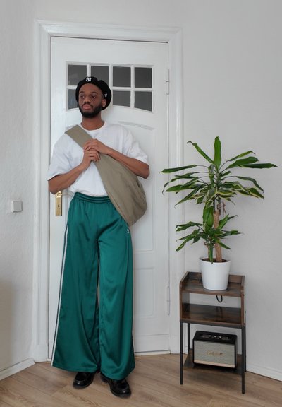Pantalones anchos de satén verde con rayas laterales blancas, camiseta blanca de gran tamaño, bolso cruzado beige, boina negra, planta de interior, estantería de madera.