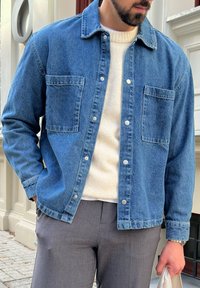 Blå denimskjorta med två framfickor, tryckknappar, avslappnad passform, lager över en beige sweater, i kombination med grå byxor.