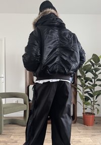 Doudoune noire avec une capuche bordée de fourrure, tissu texturé, coutures froncées et poignets côtelés. Associée à un pantalon noir ample.