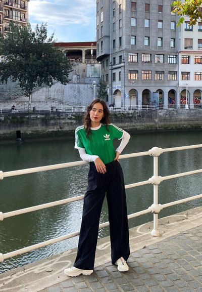 Camiseta verde de Adidas con rayas blancas sobre una camisa blanca de mangas largas, combinada con pantalones anchos de rayas negras y zapatillas blancas.