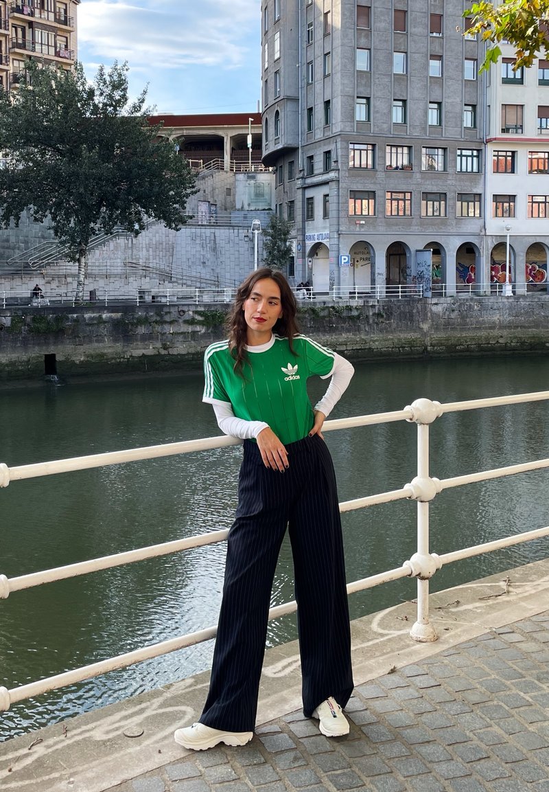 Πράσινο T-shirt Adidas με λευκές ρίγες, φορεμένο πάνω από λευκή μακρυμάνικη μπλούζα, συνδυασμένο με μαύρο παντελόνι με ρίγες και λευκά sneakers.