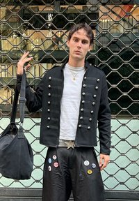 Veste noire de style militaire avec des boutons argentés, portée sur une chemise blanche. Pantalon noir ample orné de divers boutons colorés et d'une ceinture avec une étoile en argent.