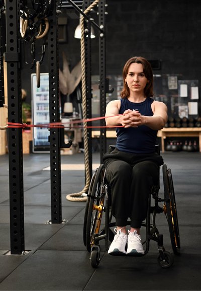 Mujer en silla de ruedas haciendo ejercicio con una banda de resistencia estirada entre las manos y el equipo de gimnasio en un centro de fitness.