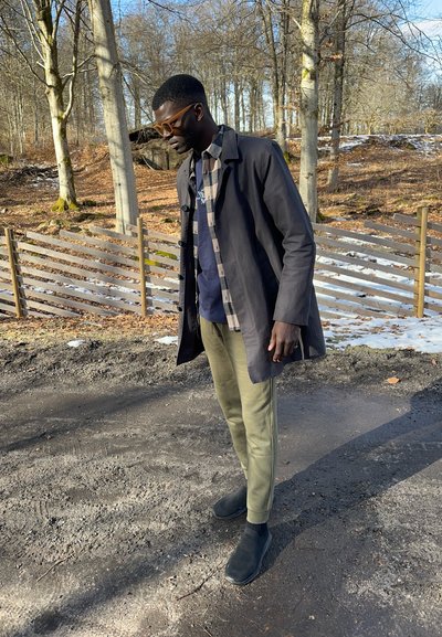 Chaqueta ligera negra sobre una camisa azul con patrón a cuadros. Pantalones verde olivo con rayas laterales y zapatos negros de tipo slip-on. Entorno al aire libre.