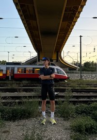 En atlet iförd en mörk tröja och svarta shorts står på järnvägsspåren, med en gul bro ovanför och ett tåg som blåser förbi.
