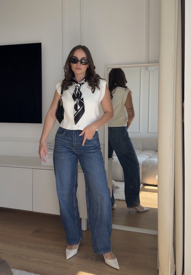 Femme portant des lunettes de soleil, un gilet blanc sans manches, une écharpe noire à motifs, un jean bleu et des talons blancs, debout à l'intérieur devant un miroir.
