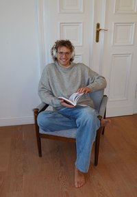 Une personne est assise sur une chaise en bois, portant un pull gris douillet et un jean bleu clair, lisant un livre et portant un casque blanc.