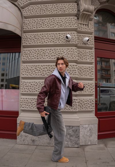 Chaqueta de cuero marrón sobre sudadera gris con capucha, vaqueros anchos grises y botas color canela. Bolsa negra con correa sostenida en la mano, de pie cerca de una pared texturizada.