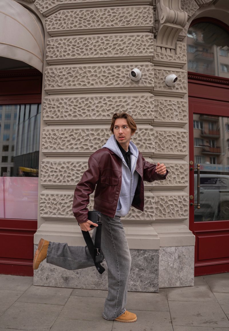 Vīna sarkanas ādas jaka pār pelēku kapuci, pelēkas platās bikses un bēšas zābaki. Melna soma ar siksnu rokā, stāvot pie teksturētas sienas.