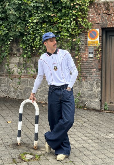 Polo blanco de manga larga con cuello negro y acentos rojos, combinado con jeans de mezclilla anchos azules y zapatos beige. Apoyado en un poste con rayas blancas.