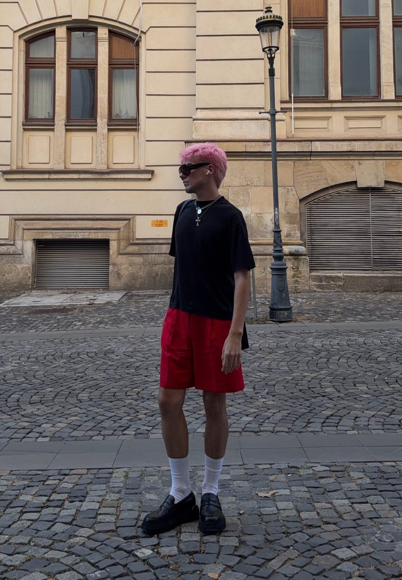 Personne aux cheveux roses portant une chemise noire, un short rouge, des chaussettes blanches et des chaussures noires, debout sur une rue pavée près d’un bâtiment beige et d’un lampadaire.