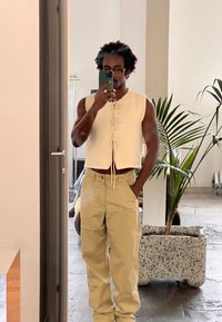 Top in maglia bianco senza maniche con dettaglio di nodo frontale, abbinato a pantaloni beige a gamba larga. Sullo sfondo si vede una pianta in vaso e un interno moderno.