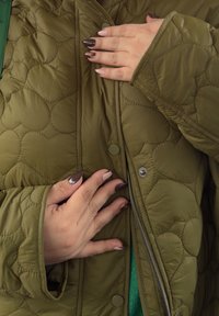 Tikkitakki oliivinvihreänä, jossa on etuvetoketju ja nepparikiinnitys. Kädet takin päällä, esittelemässä kankaan tekstuurin yksityiskohtia.