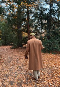 Long manteau en laine marron porté sur un pantalon clair, marchant sur un sentier couvert de feuilles avec des arbres en arrière-plan, sous un ciel dégagé.