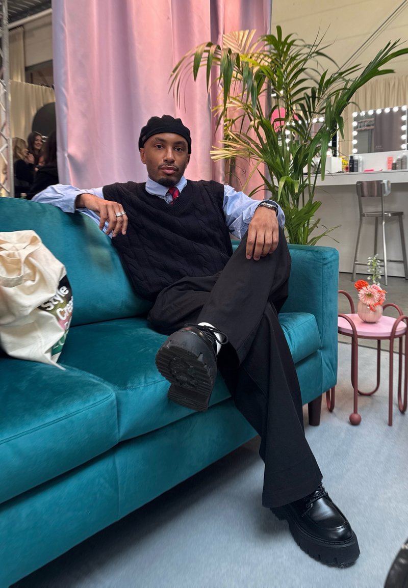 Veste sans manches en maille torsadée bleu marine portée sur une chemise bleu clair, pantalon noir et chaussures noires épaisses, assis sur un canapé bleu sarcelle avec un fond rose.