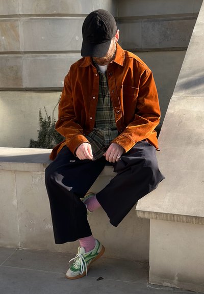 Hombre con gorra negra, chaqueta naranja, camisa de cuadros, pantalones anchos negros, calcetines rosas y zapatillas verdes sentado en un borde de concreto, mirando hacia abajo, con las manos cerca del regazo.