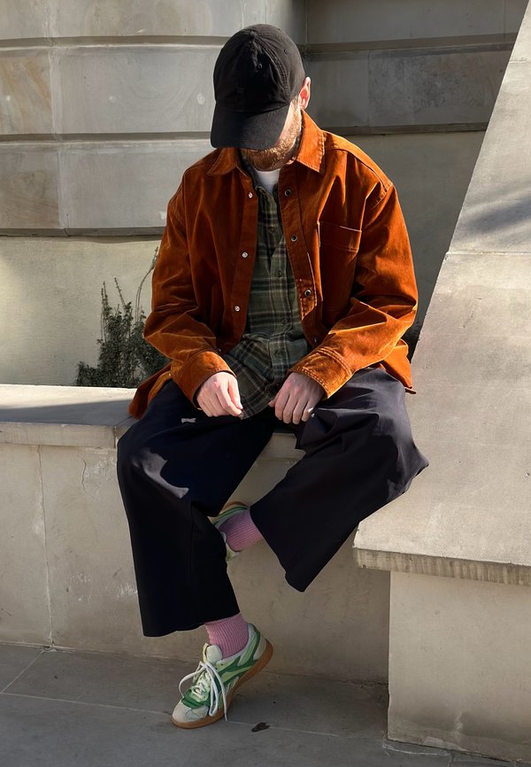 Fekete sapkás, narancssárga dzsekis, kockás inges férfi fekete, bő nadrágban, rózsaszín zokniban és zöld tornacipőben, aki egy betonperemen ül, lefelé néz, kezei az öléhez közel.