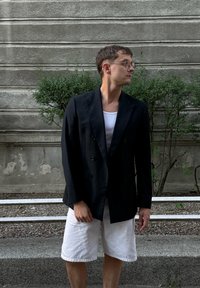 Juodas dvigubai užsegamas švarkas su viena kišene, dėvimas ant baltos tamprės, derinamas su baltais lino šortais, prieš tekstūruotą pilką foną.