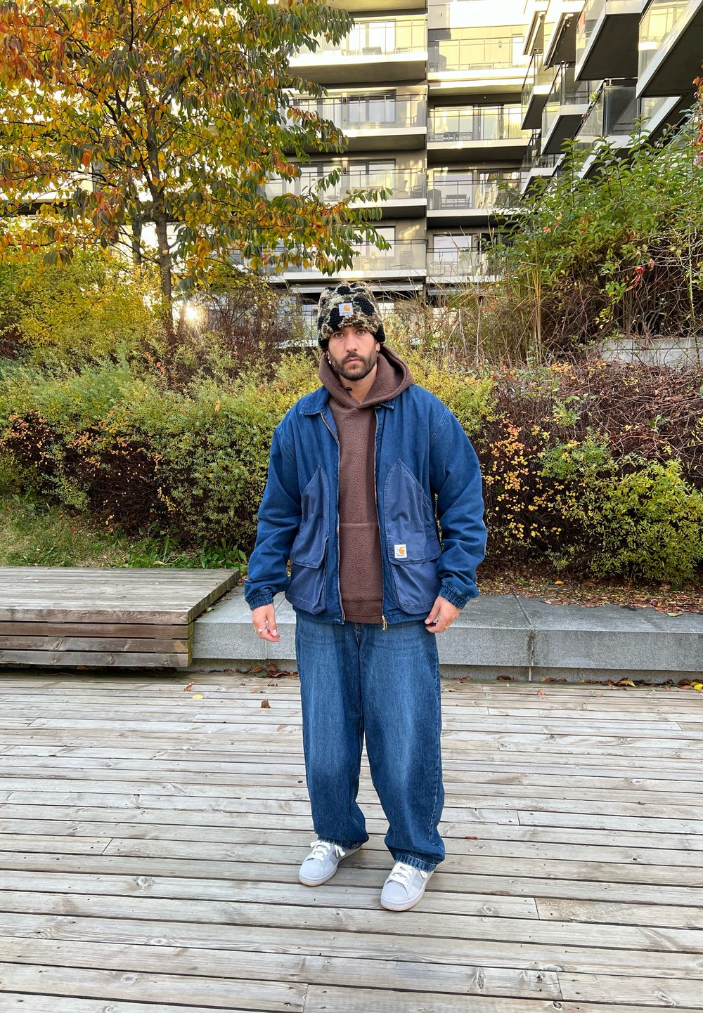 Giacca in denim con tasche a contrasto, felpa marrone sotto, jeans blu larghi e berretto lavorato a maglia con motivo. Sneakers bianche completano l'outfit.