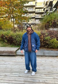 Giacca in denim con tasche a contrasto, felpa marrone sotto, jeans blu larghi e berretto lavorato a maglia con motivo. Sneakers bianche completano l'outfit.