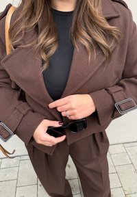 Καφέ καμπαρδίνα από μίγμα μαλλιού με υπερμεγέθη γιακά και ζώνες στα μανίκια, φορεμένη πάνω από μια μαύρη ριχτή μπλούζα. Μαύρα γυαλιά ηλίου κρατημένα σε ένα χέρι.