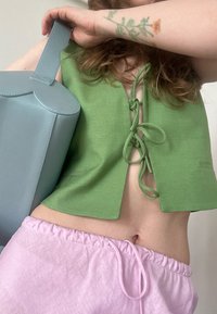 Top verde cropped com laços frontais, combinado com calções cor-de-rosa com cordão. Uma mala azul-clara com parte superior arredondada é segurada em uma mão.