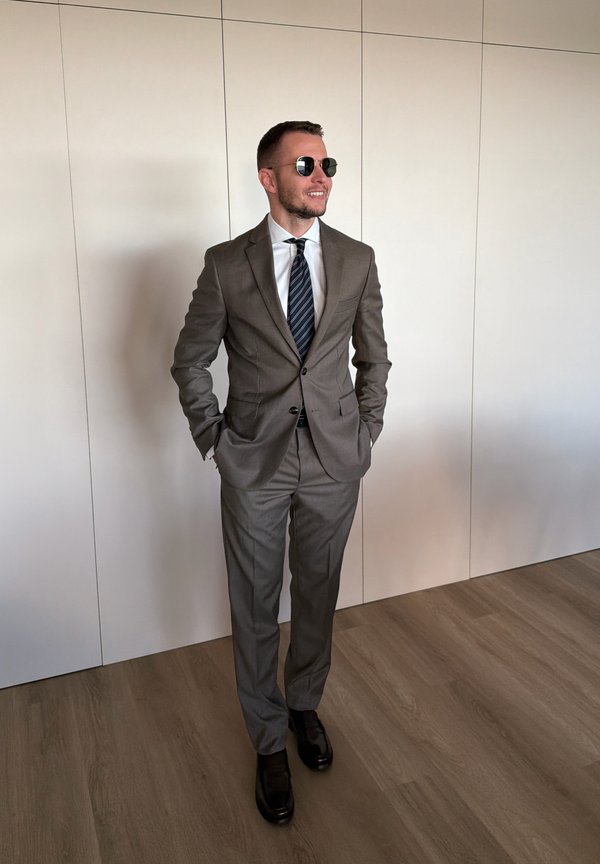 Hombre con traje gris, camisa blanca, corbata a rayas, zapatos negros y gafas de sol, de pie con las manos en los bolsillos contra una pared blanca y suelo de madera.