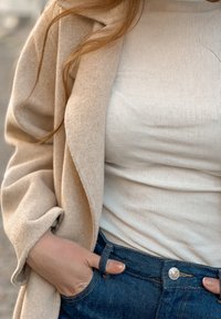 Un vêtement d'extérieur en mélange de laine beige tombe sur un débardeur blanc côtelé, associé à un jean denim bleu avec des accessoires argentés et une silhouette ajustée.